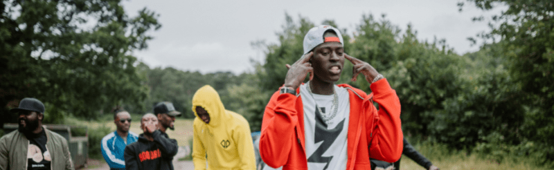 Man in red jacket performs energetically outdoors with a group of men in hoodies and caps standing behind him.