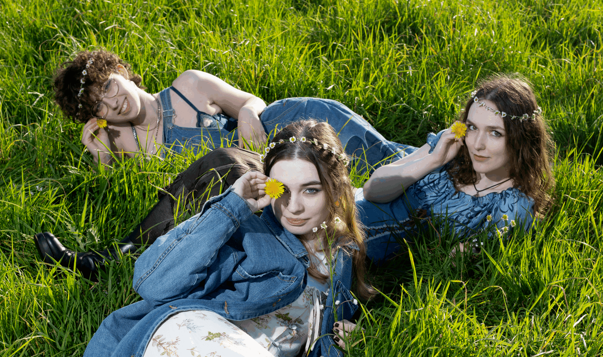 Three women in daisy chain crowns lie in tall grass, each holding a yellow flower to their face in a playful, summery pose.