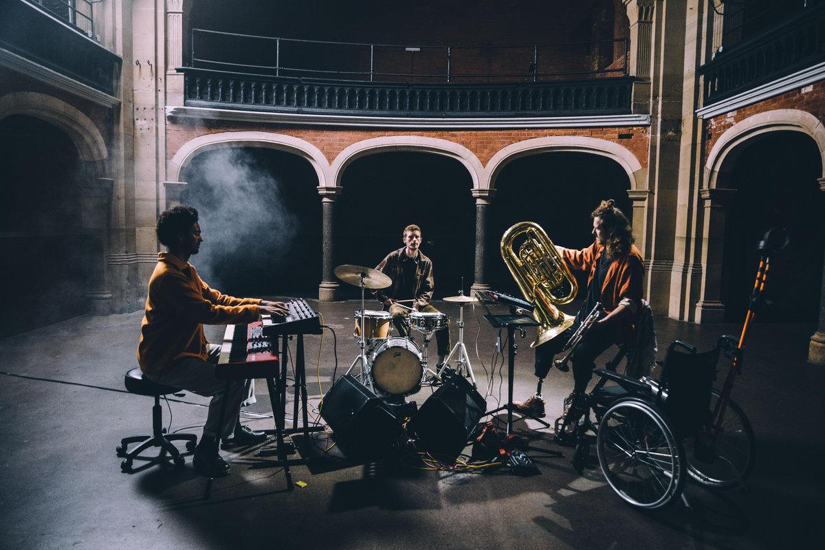 Three musicians play keyboard, drums, and tuba in an atmospheric hall with arched brickwork and smoke drifting in the background.